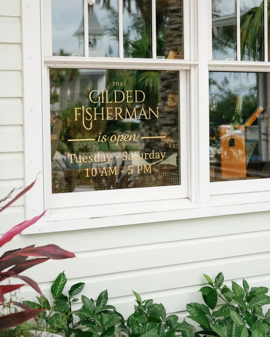 A front window in gilt lettering — The Gilded Fisherman is open Tuesday to Saturday, 10 AM to 5 PM — over a bed of red-leaved plants