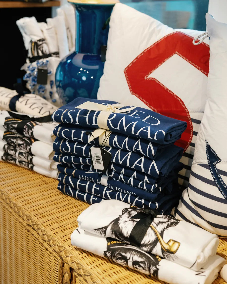 A stack of folded navy tea towels lettered The Gilded Fisherman beside a white sailcloth pillow with a red racing number