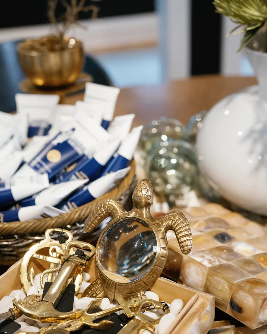 A tray of brass treasures on white pebbles: a sextant, a magnifying glass, a brass sea turtle and navy-wrapped gifts