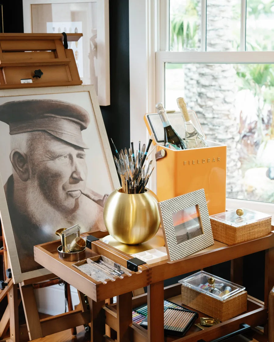 A charcoal captain's portrait on an easel beside a brass vase of brushes and a small framed sunset, on a wooden artist's cart