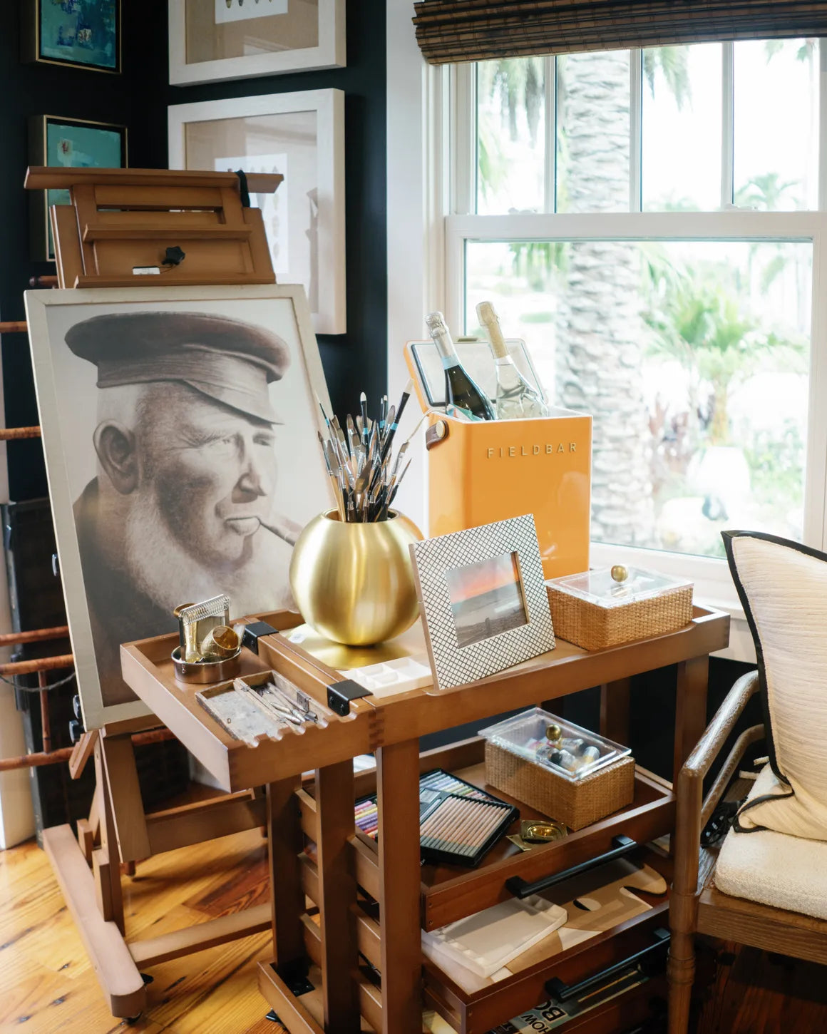 Inside the shop: a charcoal portrait of a pipe-smoking sea captain on a painter's easel, a brass vase of paintbrushes and framed pictures on the artist's cart beside it