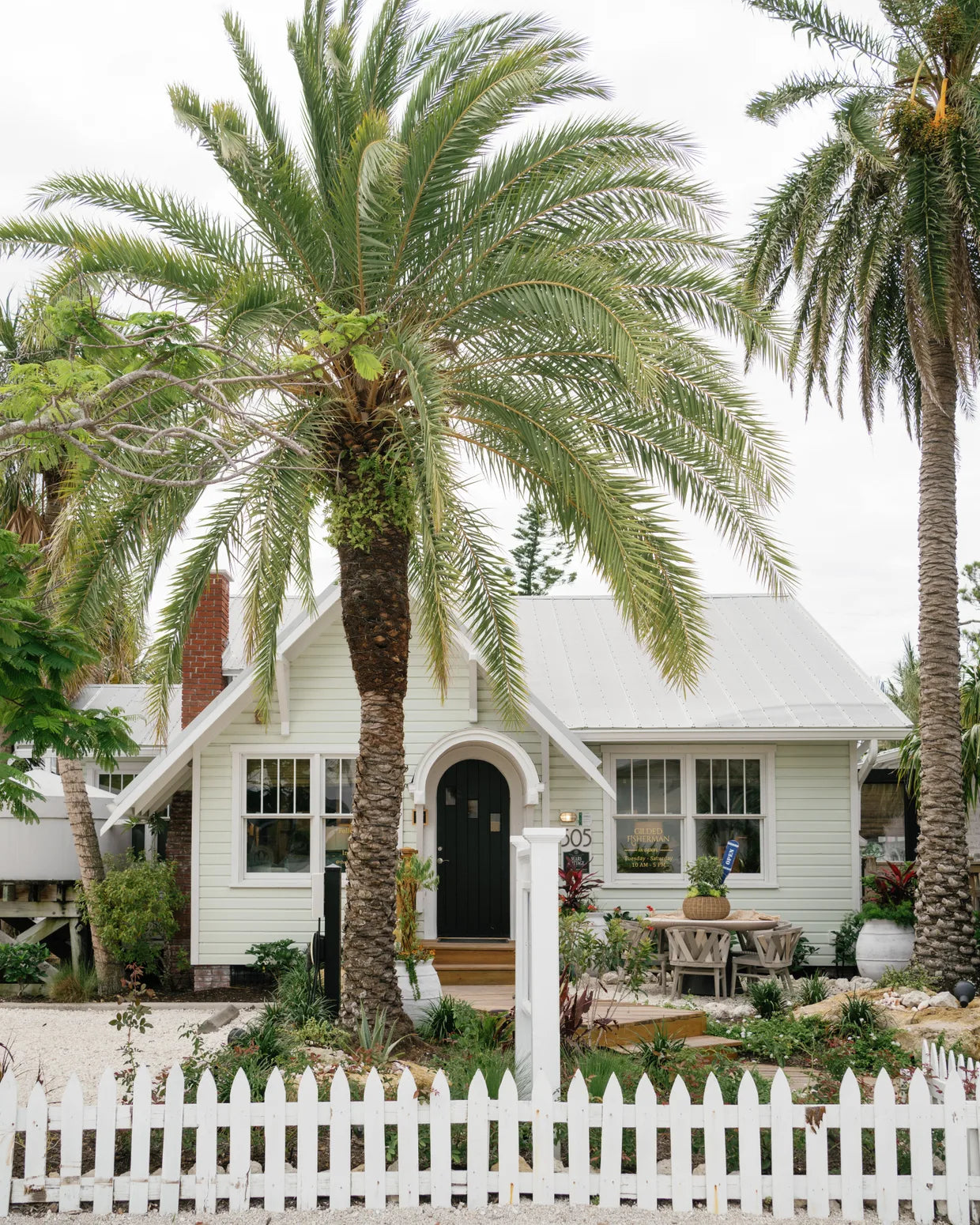 The Gilded Fisherman storefront: a pale-green island cottage with a black arched door and metal roof, framed by tall date palms behind a white picket fence