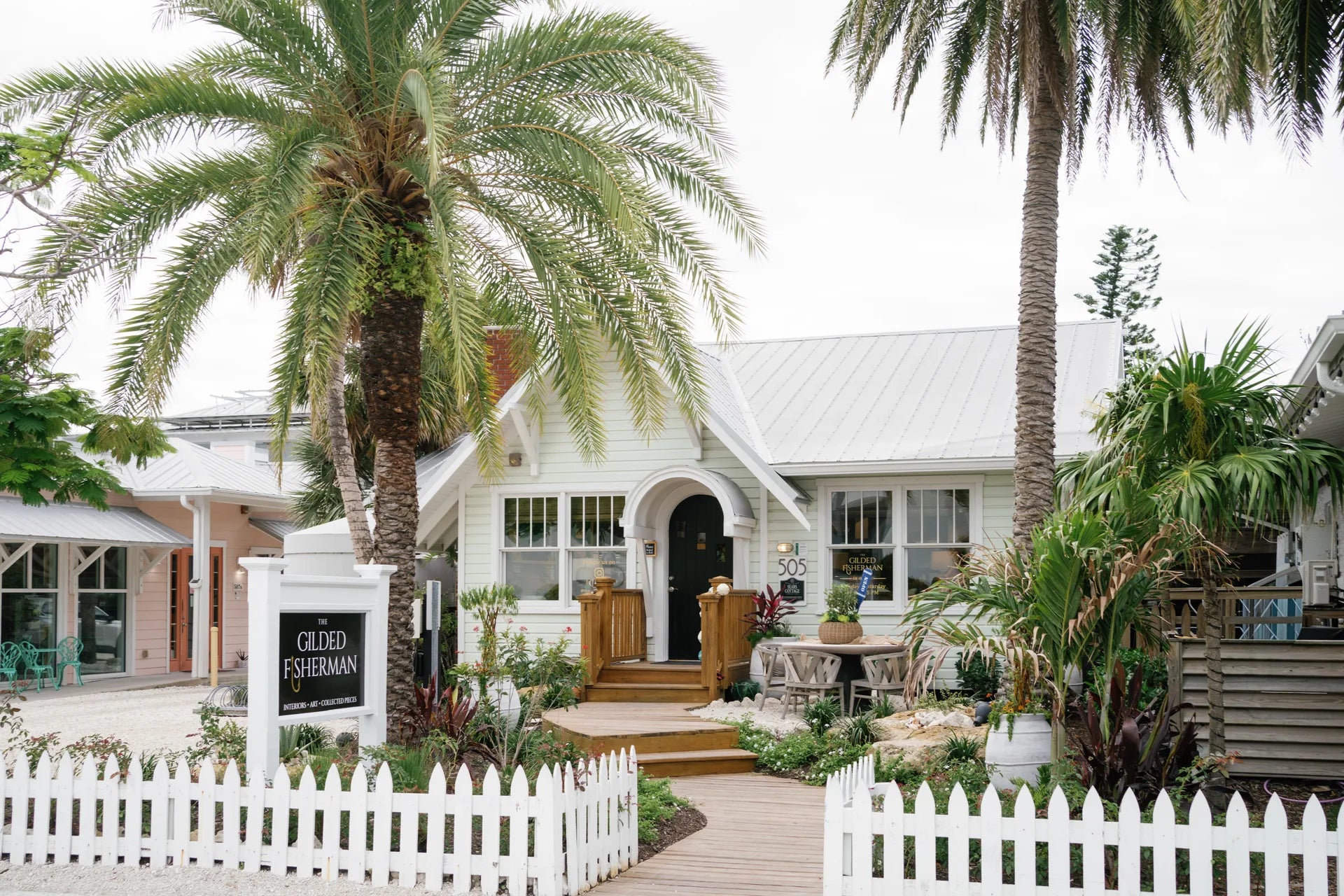 The Gilded Fisherman storefront on Pine Avenue: a pale-green island cottage with its gilt-lettered sign, framed by date palms and a white picket fence